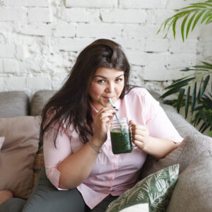 Eine junge plus size Frau sitzt gemütlich auf einem Sofa und trinkt einen grünen Smoothie aus einem Glas mit einem Strohhalm.