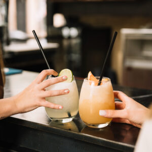 Eine Nahaufnahme von zwei Personen, die miteinander anstoßen. Sie halten jeweils ein Glass mit fruchigen Cocktails in der Hand.