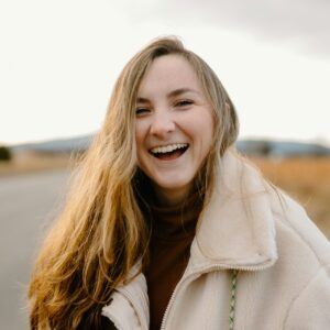 Eine junge Frau steht draußen in der Natur und lacht herzhaft in die Kamera.