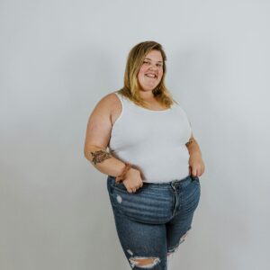 Eine slebstbewusste plus size Frau posiert lächelnd im Stehen vor einer weißen Studiowand.