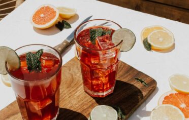 Zwei Gläser mit einem roten Cocktail befüllt stehen auf einem Holzbrett. Sie sind garniert mit Limetten und Orangenscheiben und frischer Minze.