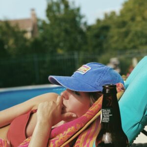 Eine junge Frau liegt auf einer Poolliege und sonnt sich. Neben ihr, im Bildvordergrund, steht eine Flasche Bier.