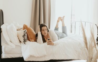 Eine junge Frau liegt auf einem weißen Bett und schaut auf ihr Smartphone.
