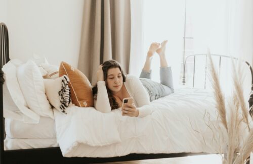 Eine junge Frau liegt auf einem weißen Bett und schaut auf ihr Smartphone.