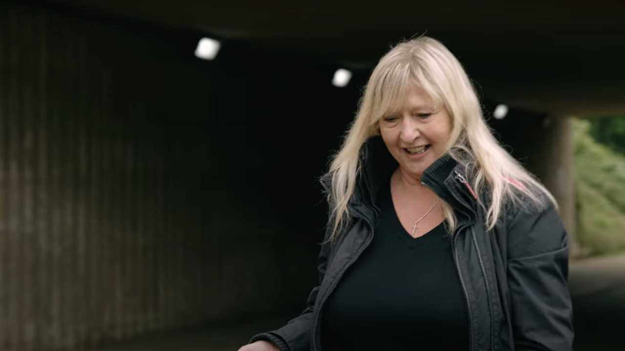 Woman walking outdoors in black jacket, smiling under a tunnel.
