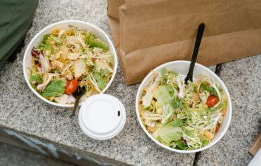 Two takeaway salad bowls with chicken, lettuce, tomatoes, and plastic forks on a granite surface.