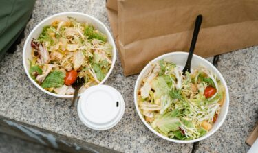Two takeaway salad bowls with chicken, lettuce, tomatoes, and plastic forks on a granite surface.