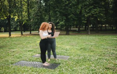 Two women standing on yoga mats in a green park, looking at a laptop, planning a workout session.