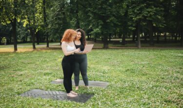 Two women standing on yoga mats in a green park, looking at a laptop, planning a workout session.