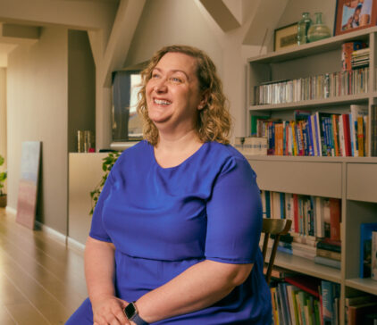 Smiling woman in a blue dress sitting in a living room with bookshelves in the background.