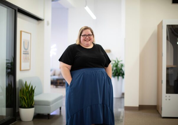 A smiling plus-size woman with glasses, wearing a black top and a blue skirt, stands confidently in a modern office space with plants, framed artwork, and seating in the background.