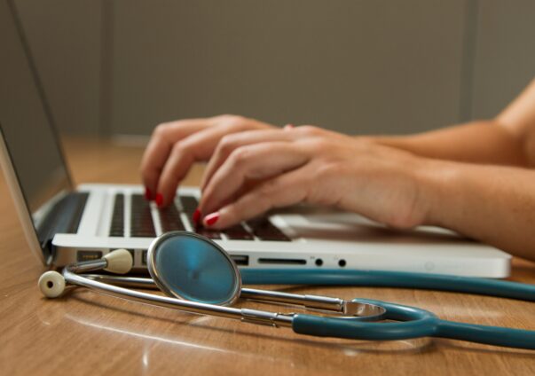 Hands typing on a laptop next to a stethoscope on a wooden desk.