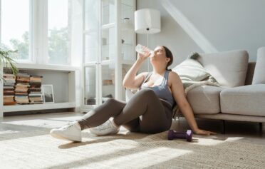 Woman in workout clothes sitting on the floor of a bright living room, drinking water after exercising, with a dumbbell nearby.