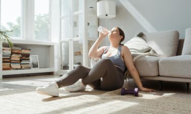 Woman in workout clothes sitting on the floor of a bright living room, drinking water after exercising, with a dumbbell nearby.