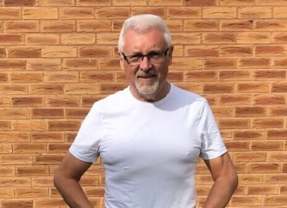 Confident older man in white t-shirt and jeans standing outside by a brick wall.