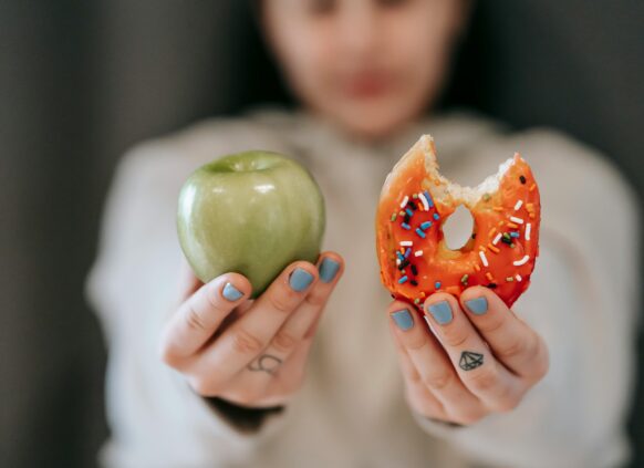 Person holding a green apple in one hand and a bitten donut with sprinkles in the other, symbolizing food choice.