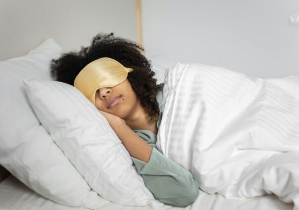 Young woman sleeping peacefully in bed with a gold eye mask and white bedding.