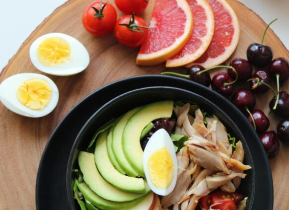 Healthy meal with avocado, chicken, hard-boiled eggs, tomatoes, grapefruit slices, and cherries on a wooden board.