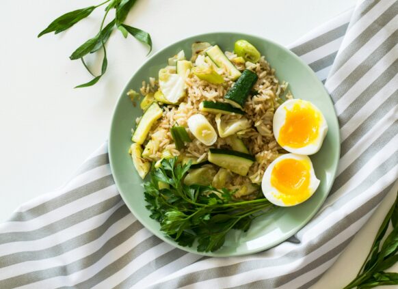 Plate of rice with zucchini, leeks, soft-boiled eggs, and fresh herbs on a striped cloth background.