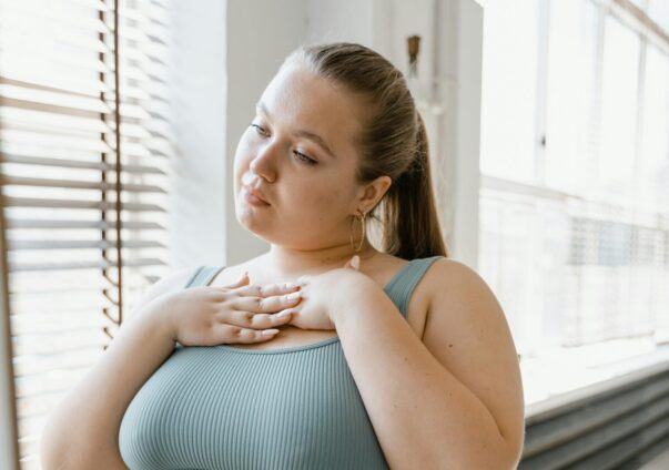 Young woman with her hands on her chest, standing by a window and reflecting with a calm expression.