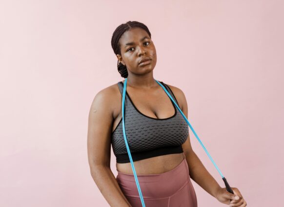 Young woman in activewear holding a blue jump rope with a neutral expression against a pink background