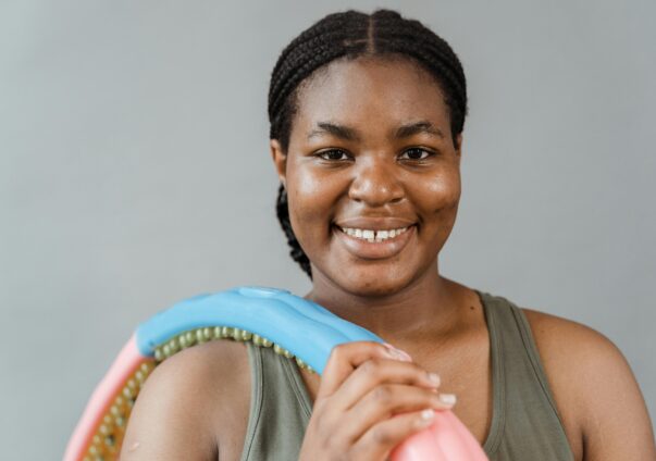 Smiling woman in sportswear holding a colorful fitness hoop against a gray background.