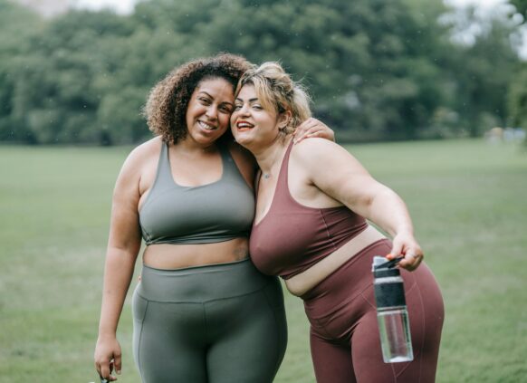 Two plus-size women in activewear smiling and hugging after a workout in the park