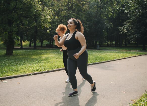 Two women in activewear jogging on a park path surrounded by greenery
