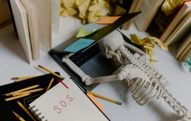 Skeleton leaning over a laptop surrounded by books, notes, and post-its, symbolizing study burnout.