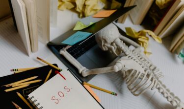 Skeleton leaning over a laptop surrounded by books, notes, and post-its, symbolizing study burnout.