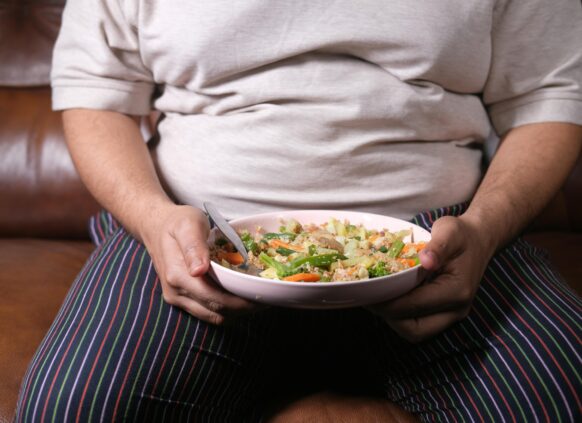 Overweight man in pajamas holding a bowl of mixed vegetables and grains while sitting on a couch.