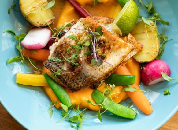 Grilled fish fillet served with colorful vegetables, mashed sweet potatoes, and microgreens on a blue plate