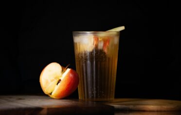 Glass of apple juice with ice and apple slices, next to a fresh red apple on a wooden board.