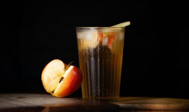 Glass of apple juice with ice and apple slices, next to a fresh red apple on a wooden board.