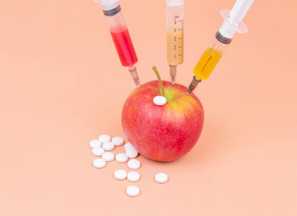 Apple injected with colorful syringes surrounded by white pills on a peach background