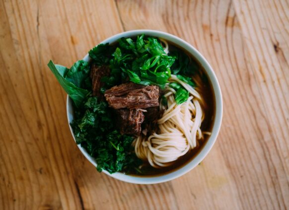 Bowl of noodle soup with beef, herbs, and broth on wooden table