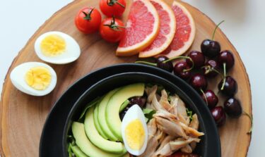 Healthy lunch bowl with avocado, egg, chicken, tomatoes, cherries, and grapefruit slices on a wooden plate