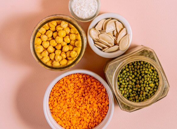 Assorted bowls of lentils, chickpeas, mung beans, pumpkin seeds, and shredded coconut on a pink background