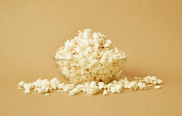 Glass bowl of popcorn with scattered kernels on beige background