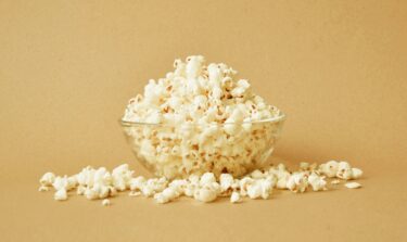Glass bowl of popcorn with scattered kernels on beige background