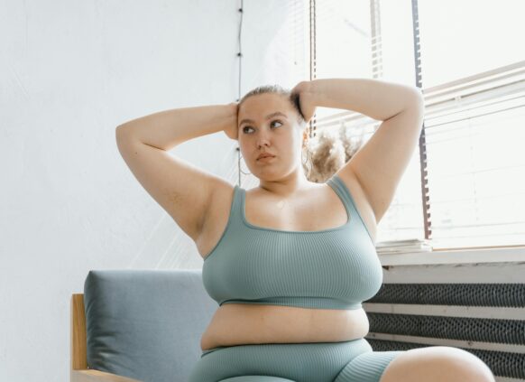 Plus-size woman in activewear sitting indoors with hands behind head, lit by window light