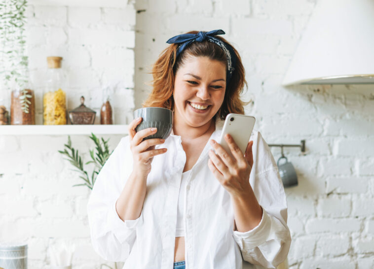 Plus-sized woman happily using her phone in the kitchen