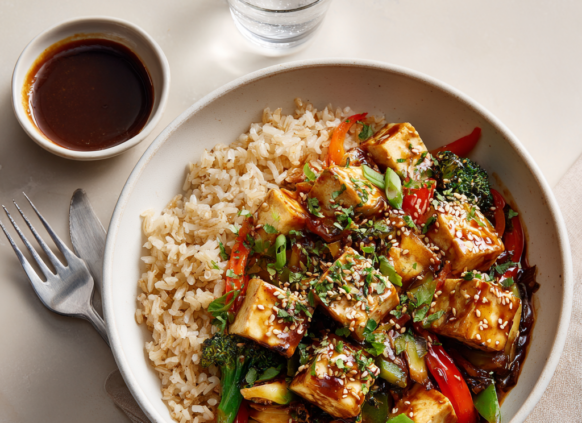 Bowl of tofu and vegetable stir fry with brown rice and a small bowl of dipping sauce